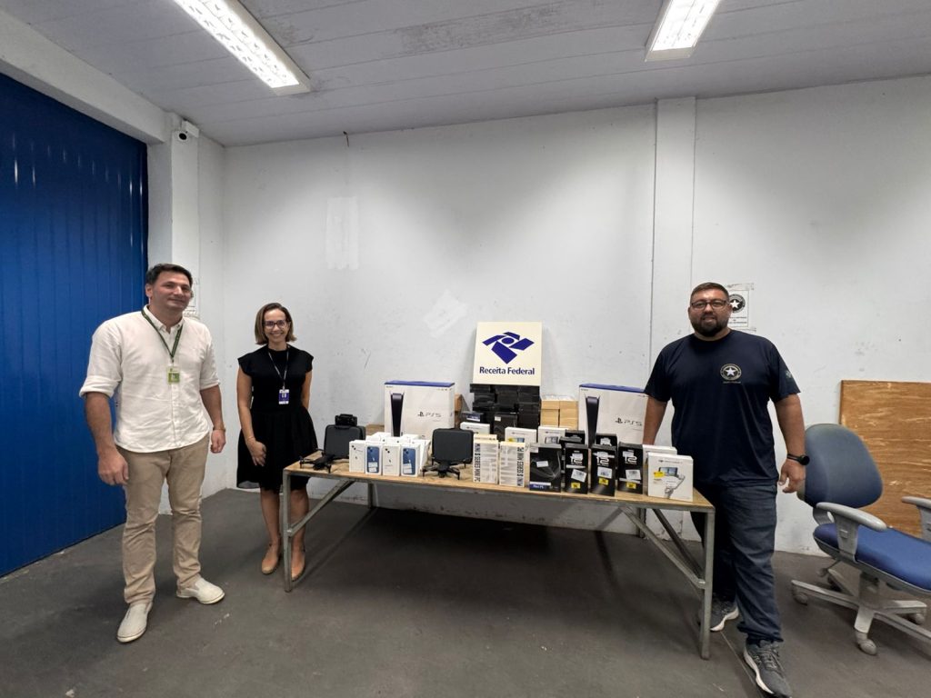 Três pessoas em pé ao redor de uma mesa com eletrônicos expostos, em uma sala interna. Sobre a mesa há caixas de videogame, celulares e outros equipamentos, com uma placa da Receita Federal ao centro.