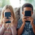 Duas crianças sentadas em um sofá seguram celulares em frente ao rosto, com a tela cobrindo os olhos.