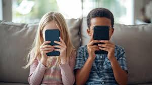 Duas crianças sentadas em um sofá seguram celulares em frente ao rosto, com a tela cobrindo os olhos.